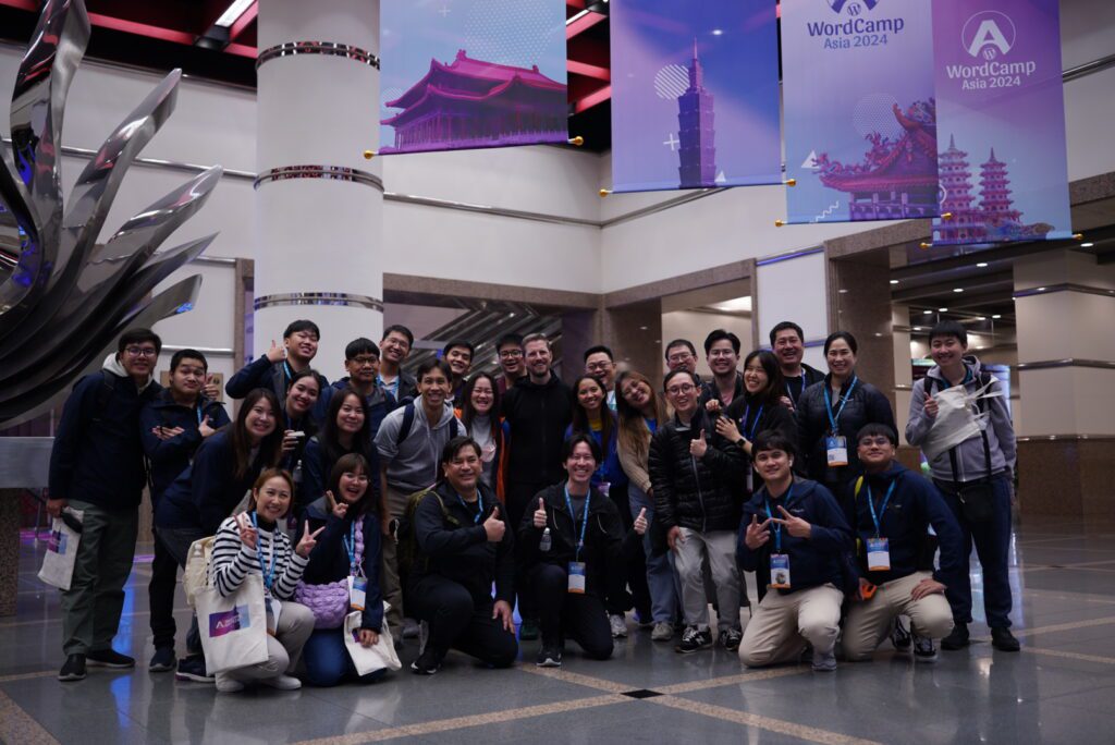 A smiling group of people crowded together for a photo, giving thumbs up and peace signs, and wearing conference lanyards.