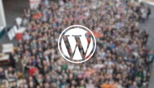 A large group of people is gathered in a blurred setting, likely at an event or conference. In the foreground, a prominent white logo of WordPress is centered, featuring a stylized "W" within a circle. The crowd appears diverse, with individuals wearing various colored shirts and engaged in conversation or posing for a photo. The background is filled with indistinct figures, creating a sense of a lively gathering.
