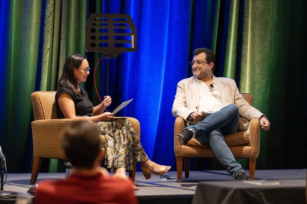 A woman and a man sit in armchairs on stage, engaged in conversation.