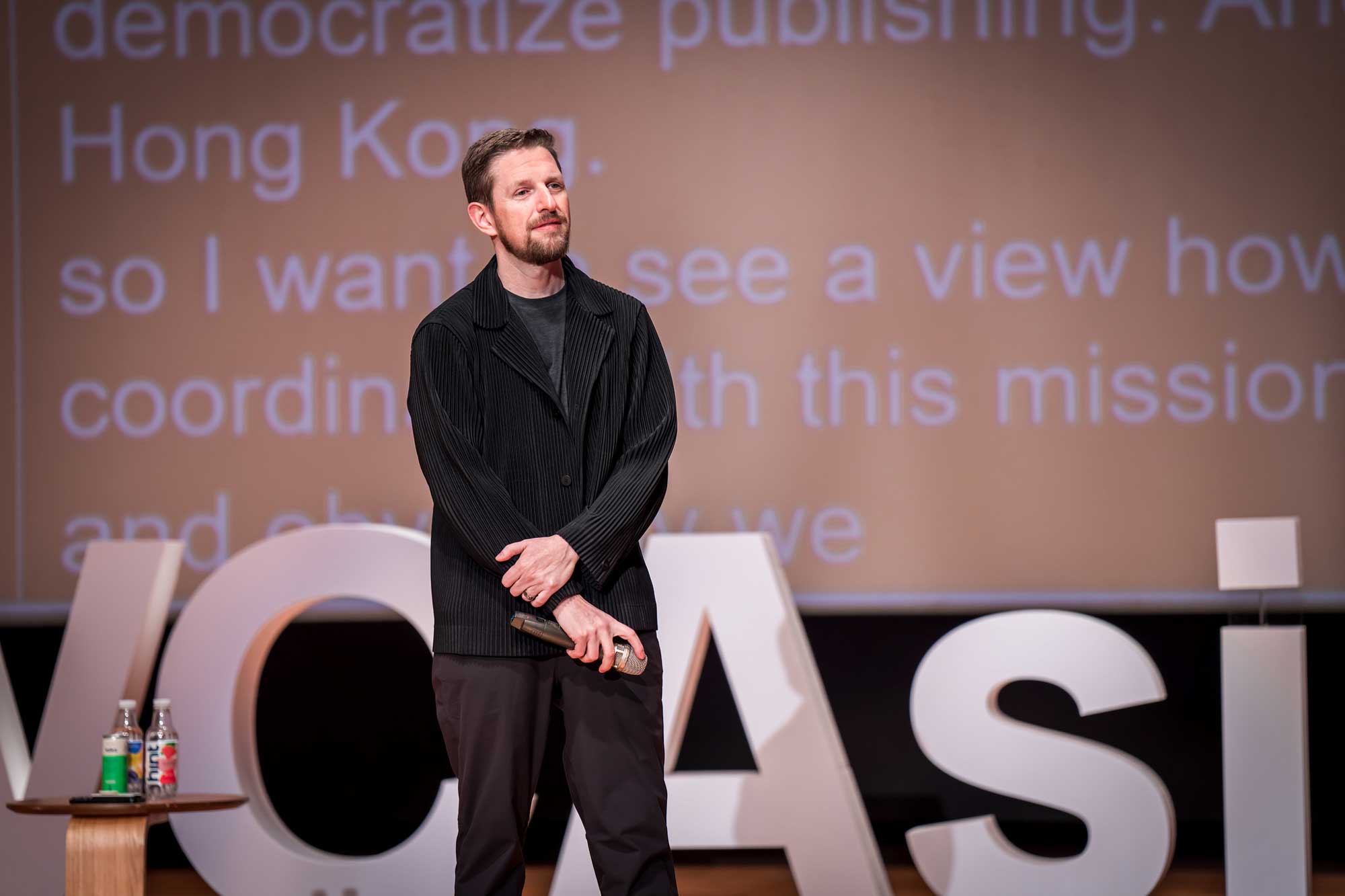 A speaker stands on stage with a microphone, addressing an audience.