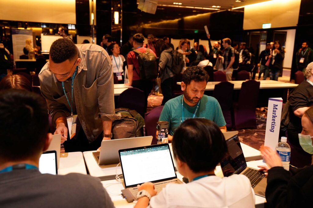 A group of people collaborating at tables with laptops in a conference setting.