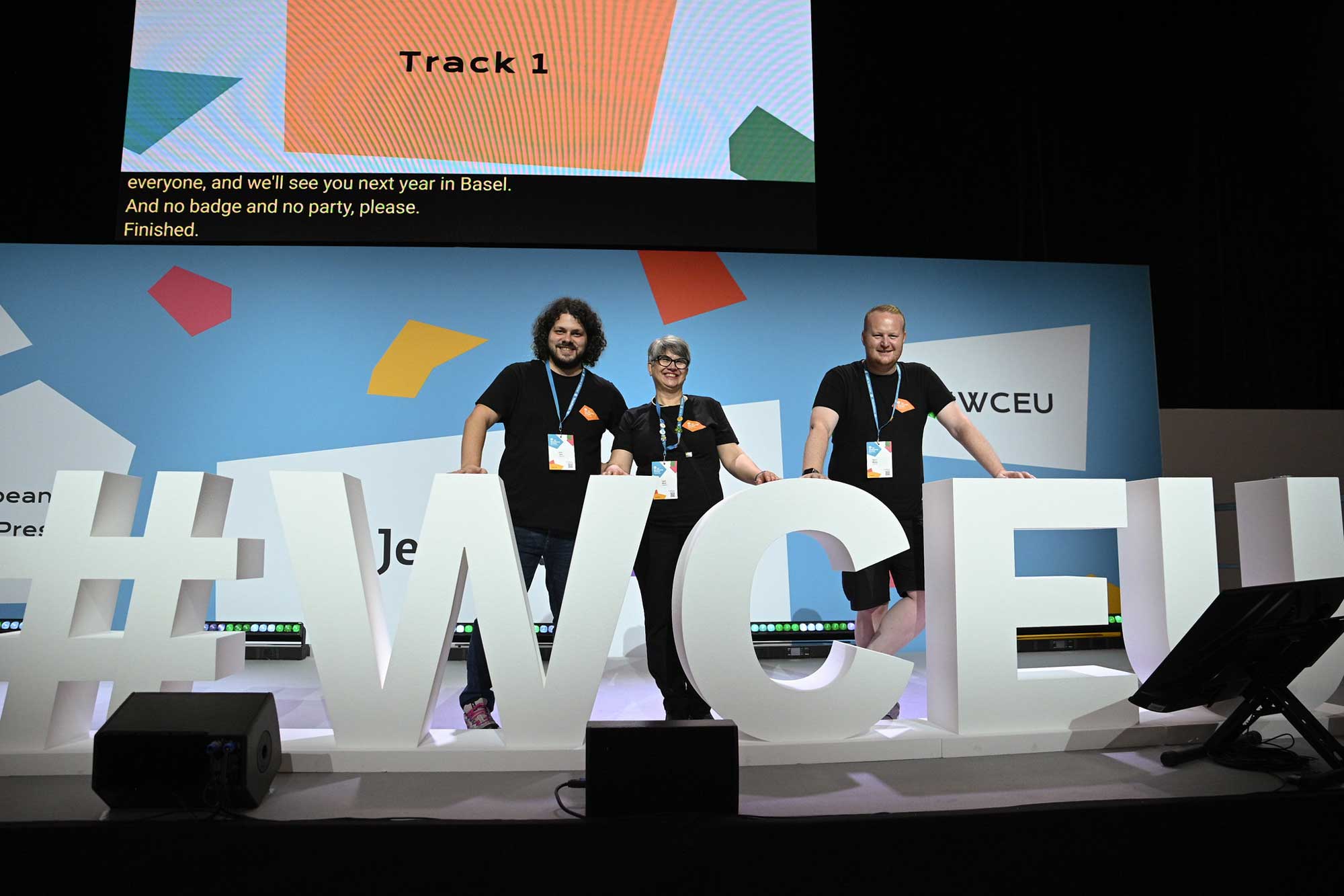 Three people stand behind a large "#WCEU" sign on stage, smiling at the camera.