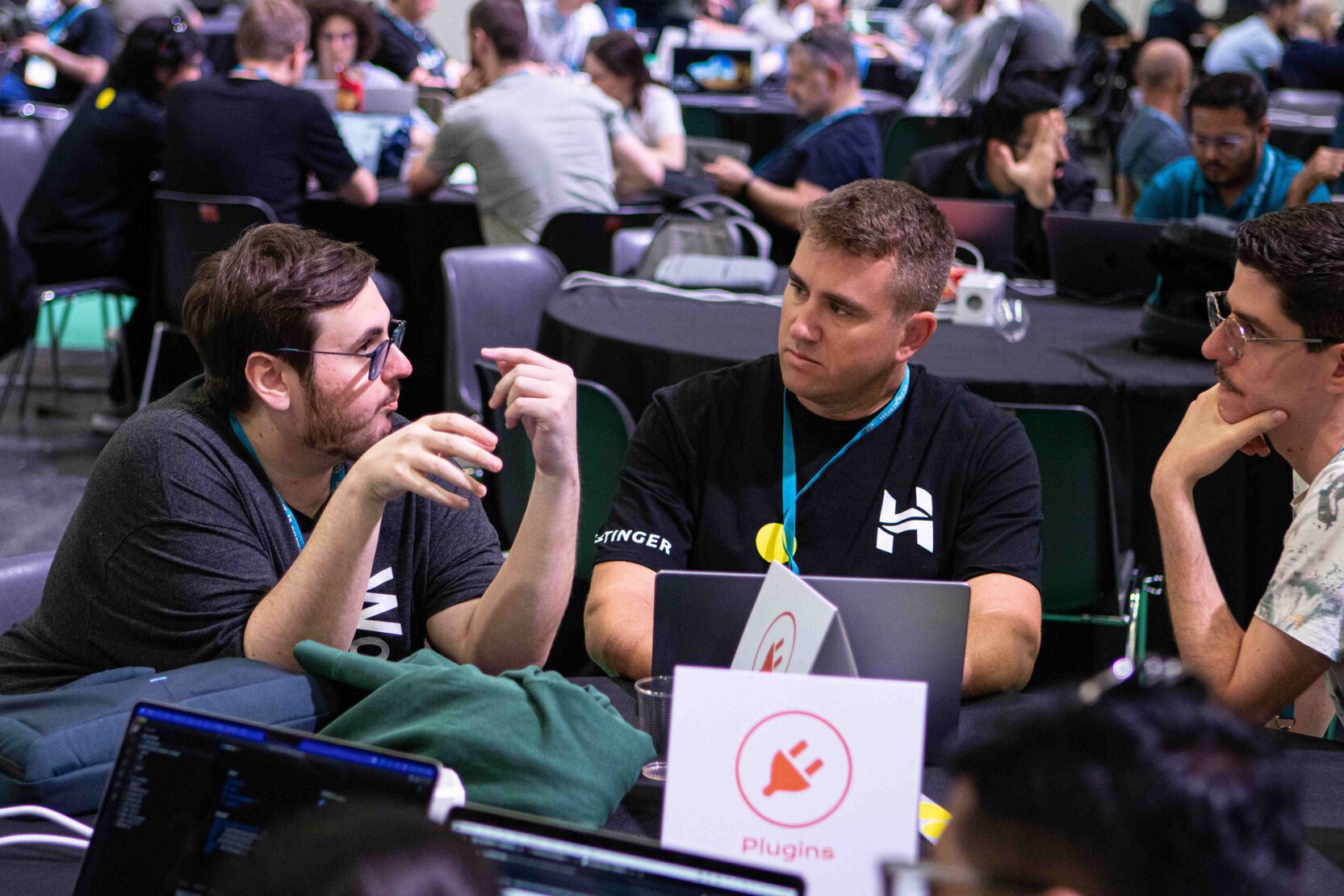 Three people engage in discussion at a conference, with laptops and tables in the background.