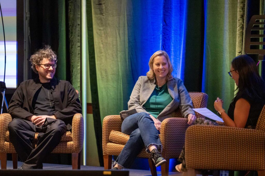 Two speakers sit on stage in armchairs, engaged in conversation at an event.