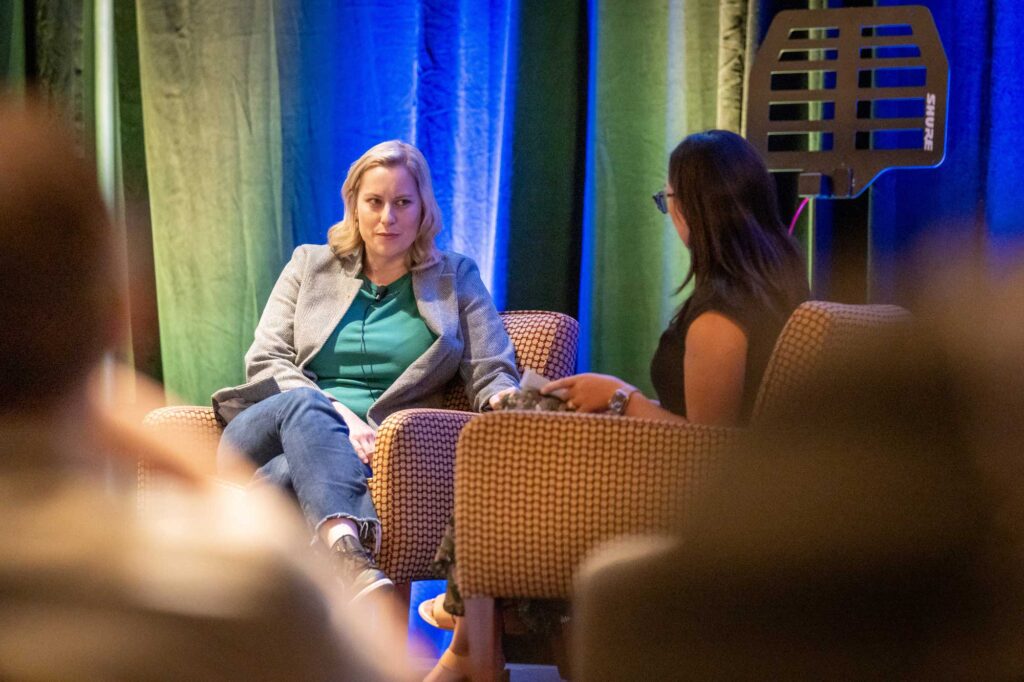 Two women sit in armchairs during a discussion, with colorful lighting in the background.