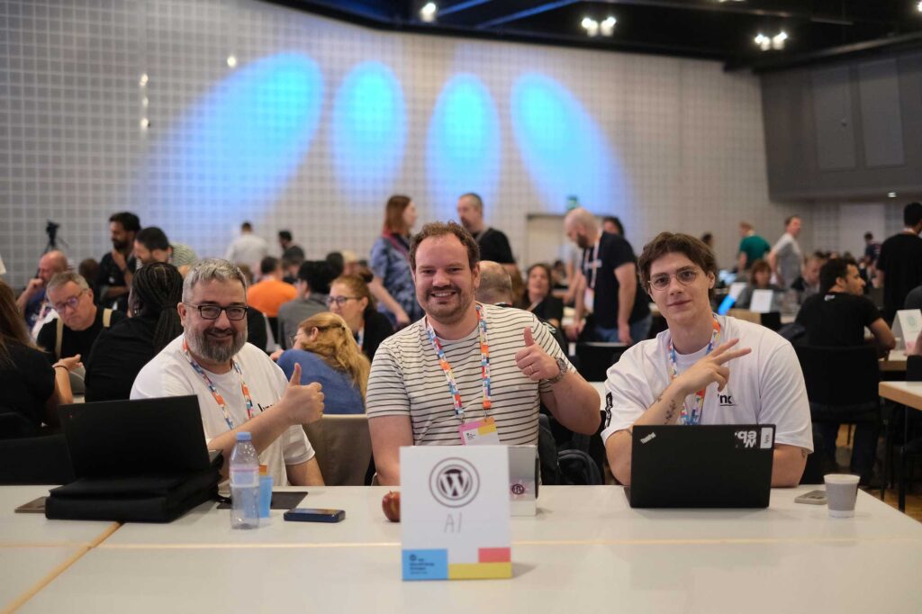 Three people sitting at a table with laptops, smiling and giving thumbs up. Background is busy.