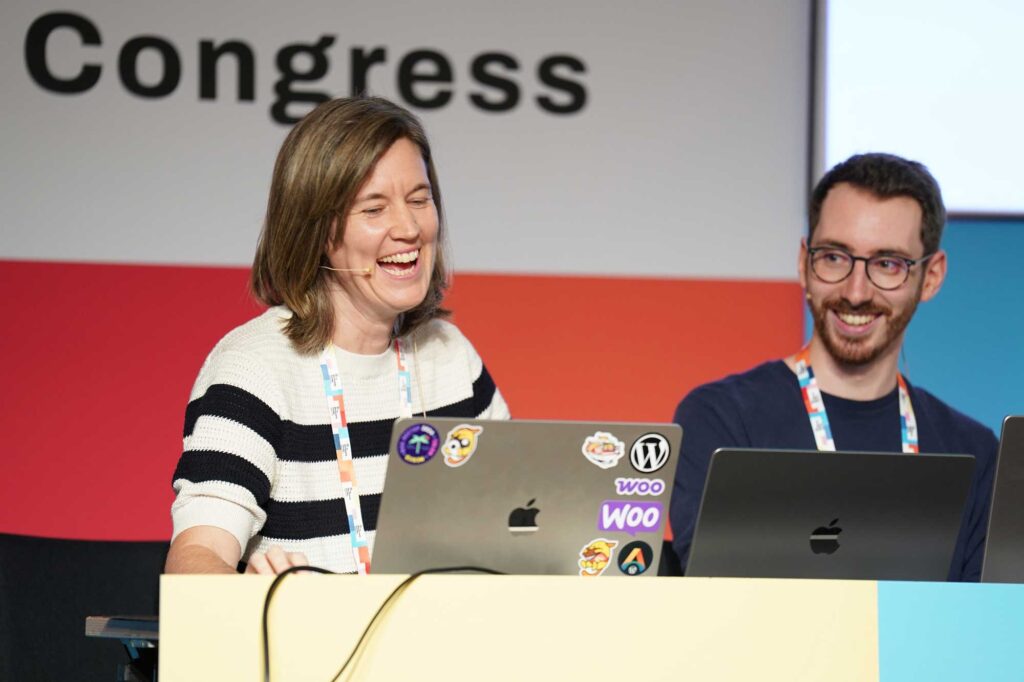 Two presenters at a conference, smiling and engaging with laptops on a stage.