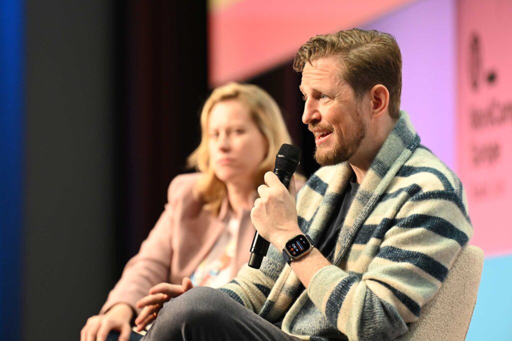 A man in a striped sweater speaks into a microphone, while a woman listens.