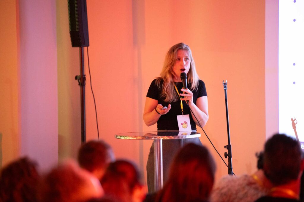 A woman speaks into a microphone at a podium, with an audience visible behind her.