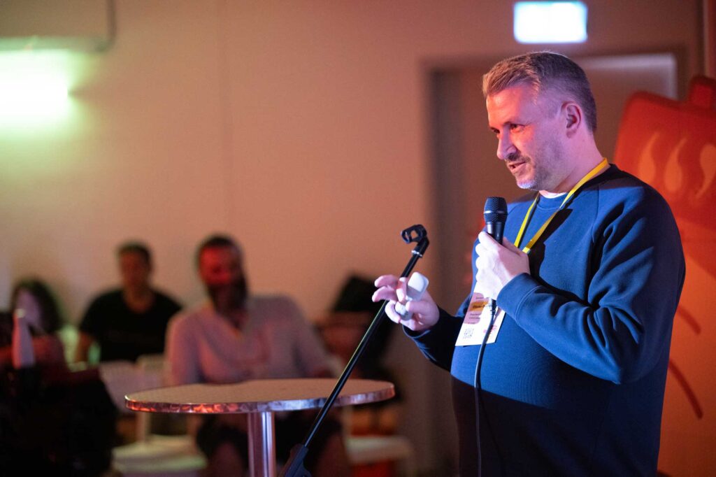 A speaker holds a microphone while addressing an audience in a dimly lit room.