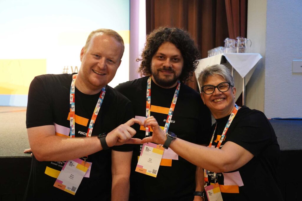 Three people in black t-shirts form a heart shape with their hands, smiling.